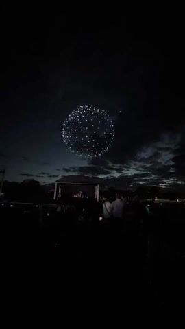 The video depicts an outdoor event at night with a crowd of people facing a brightly lit stage. Above the stage, a formation of illuminated drones creates a circular pattern in the sky. People in the crowd are holding up their phones, presumably recording the spectacle. There are some trees and a dark cloudy sky in the background. The video is dimly lit, accentuating the lights from the stage and drones.