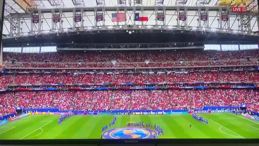 This media file is a video clip showing a wide view of a packed stadium during a soccer match. The stands are filled with spectators wearing predominantly red clothing, creating a vibrant and enthusiastic atmosphere. On the field, players in blue and red uniforms are lined up in a circle around a large circular logo depicting a globe. The field itself is a lush green, with white lines marking the boundaries. Above the stands, banners with various insignias are visible, along with the American and Texas flags, suggesting a possible location in Texas, USA, or an event with US ties. The sky is not visible, but the stadium lighting suggests it could be evening or an indoor event. The overall mood appears to be one of anticipation and excitement for the game about to begin. Some visible text includes "LIVE" in the upper right corner and "SKYWORTH" at the bottom of the screen. The clip appears to be a pre-game ceremony or introduction.