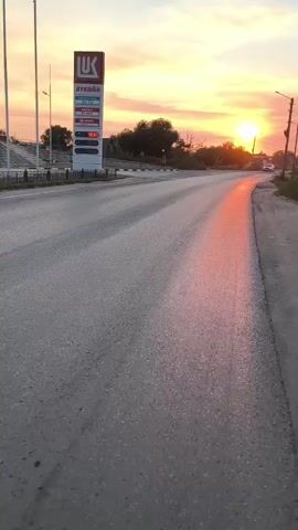 The video shows a paved road during sunset. On the left side of the road is a tall sign for a LUKOIL gas station with prices displayed. A white SUV is driving down the road, reflecting the sunset. The sky is filled with orange and pink clouds. There are trees and houses in the background. The road appears wet, with reflections of the sunset visible on the surface.