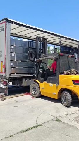 A yellow forklift, with a person in a red shirt driving, is actively unloading a semi-truck filled with what appear to be metal panels or beams. The truck's trailer is partially open, revealing stacks of the long, flat items. The forklift's mast is extended, lifting a load from the truck bed. The scene takes place outdoors on a sunny day, indicated by the bright blue sky and clear shadows. The ground is paved, suggesting a loading dock or industrial area. In the background, another truck with a blue tarp covering its load is parked, and some trees are visible.

Visible text includes "N50" on the forklift, and markings on the semi-truck trailer. The overall impression is of a routine industrial operation.
