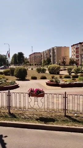 The video shows a view of a city park with apartment buildings in the background. A metal fence with decorative elements separates the park from a sidewalk and road. Flower beds with colorful flowers are visible in the park. A lamppost with two security cameras is present in the foreground, casting a shadow on the ground. The sky is clear and blue, indicating a sunny day. Trees and bushes are scattered throughout the park, contributing to the green landscape. The apartment buildings are multi-story structures with balconies and windows. The overall scene depicts a calm and orderly urban environment.