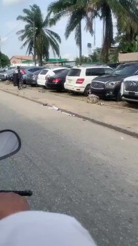 The video shows a moving perspective from a vehicle, possibly a motorcycle, driving along a road in what appears to be a tropical or subtropical location. Several cars are parked along the side of the road, near palm trees. As the vehicle advances, a gas station comes into view, identified by the green and white "AP" logo. The road shows signs of wear and tear. In the initial frames, a person dressed in a dark robe or gown, is walking along the sidewalk. A communication tower can be seen in the distance. The environment appears to be a mix of residential and commercial areas.