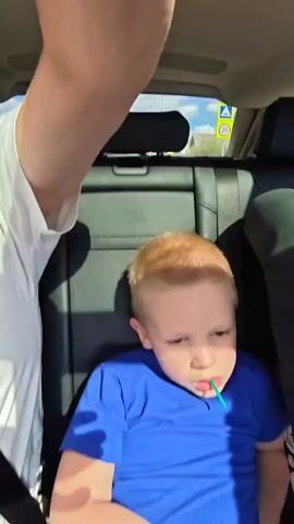 A young boy with blonde hair wearing a blue t-shirt and dark pants sits in a car and chews on a blue object. The camera then pans to a view from inside the car looking up at the sky through a sunroof. The sky is blue with white clouds and the sun is shining. Trees are visible through the sunroof.