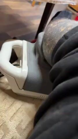 The video shows a person seated at a table, with a foot massager placed beneath their leg. The massager is primarily white and gray. The person's leg is clad in a black sock and dark gray sweatpants. The leg features visible tattoos. Above the table are numerous glass cups, and a television can be seen in the background. The table is covered with a patterned tablecloth. The camera is angled downwards, close to the massager and the person's leg.