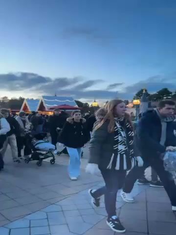 The video shows a bustling scene at what appears to be a Disney theme park at dusk. A crowd of people, many wearing winter clothing, stroll along a brick-paved path. Some people are pushing strollers. In the background is a whimsical, colorful structure that looks like a miniature castle or village, decorated with flags and lights. The sky is a mix of blue and orange, indicating it's early evening.