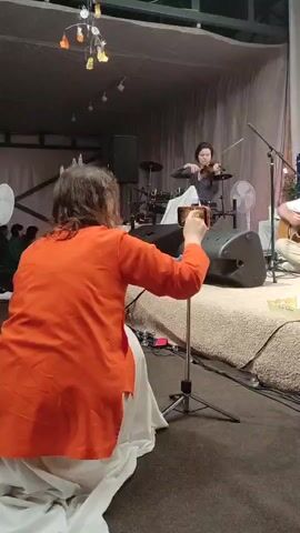 A woman in an orange shirt films a woman playing the violin on a stage. The woman playing the violin is wearing a sparkly black top and has her hair pulled back. She is standing in front of a drum set and some speakers. There is also a person playing guitar to the right of the frame, partially obscured by speakers. The camera recording the violinist is on a tripod in front of the woman in the orange shirt. The environment appears to be an indoor venue with curtains in the background and lights hanging from the ceiling.