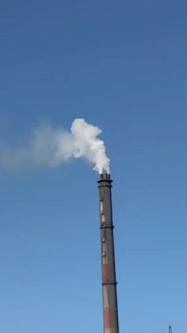 A tall industrial smokestack stands against a clear blue sky, emitting a plume of white smoke. The smokestack is made of metal and has several sections with visible bolts or seams. Red and gray bands are present on the lower portion of the smokestack. The smoke billows upwards and drifts to the left, dissipating into the atmosphere. The scene suggests an industrial setting, possibly a factory or power plant. The time of day appears to be daytime due to the bright blue sky and sunlight. No people are visible in the frame. The weather seems clear and possibly sunny. No text is visible. The location context suggests this might be in Poland, where such industrial structures are common. The overall impression is one of industrial activity.