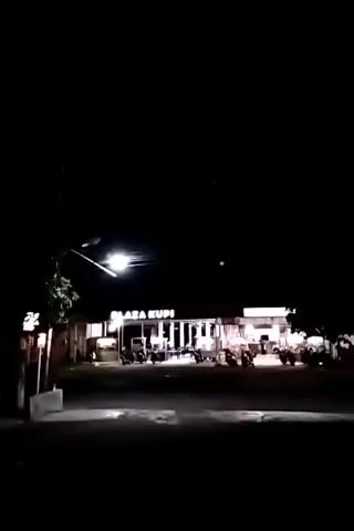 A nighttime exterior shot shows a commercial establishment labeled "BLAZA KUPI" illuminated by artificial lights. Several motorcycles are parked in front of the building. A street lamp with a bright light is visible on the left side of the frame, casting light on the foreground. The sky is dark and clear, with no visible celestial bodies. The ground appears to be asphalt or a similar paved surface. The overall scene suggests a roadside business operating after dark.