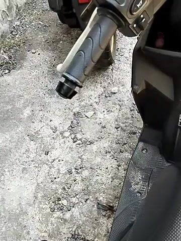 The video begins with a close-up of the handlebar and front of a black scooter. The scooter is parked on a rough, textured surface, possibly concrete or asphalt. The background is out of focus, showing what appears to be a wall and more of the scooter. The camera then pans slightly to the left, revealing another scooter parked behind the first one. This second scooter is also black and has a visible license plate that reads "DR 2990 ZR". The scene is outdoors, with a sunny sky and some greenery visible in the distance. The video continues to show the scooters and the environment, with the camera occasionally zooming in or out. The overall impression is of a casual recording, possibly taken by someone standing next to the parked scooters.