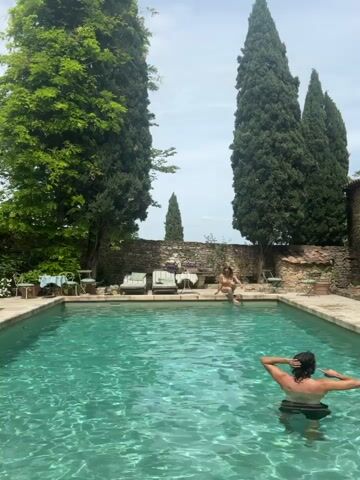 The video shows an outdoor swimming pool scene in Saint-Victor-la-Coste, France, on a bright, sunny day. Two individuals are present: one person is standing in the turquoise pool with their back to the camera, adjusting their dark hair. Another person is sitting on the edge of the pool, facing towards the person in the water, also appearing to adjust their hair or relax.

The pool area is surrounded by lush greenery, including several tall, slender cypress trees that frame the view. A stone wall runs along the far side of the pool, behind which are more trees. To the left of the sitting person, there are two lounge chairs and two small tables with chairs, suggesting a comfortable outdoor living space. The overall atmosphere is serene and leisurely.