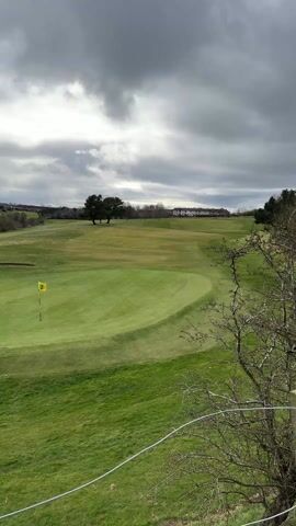 The scene is a golf course under a heavily clouded sky. The green is vibrant, and a yellow flag marks the hole's location. A sand trap is visible near the green. In the background, trees dot the landscape and a row of houses can be seen in the distance. The branches of a tree are visible in the lower right corner of the frame. Wind noise is prominent.
