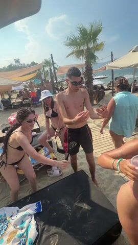A group of young adults are dancing on a sandy beach. In the foreground, two women in bikinis are dancing while holding a selfie stick. A man in swim trunks is dancing between them, and a woman in a bikini is partially visible in the lower right corner holding a drink. In the background, palm trees, beach umbrellas, lounge chairs, and other beachgoers are visible. The sky is blue with some clouds. The overall atmosphere is lively and festive.