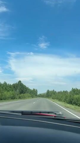 The video shows a view from inside a car traveling on a road. The road is paved with white lines marking the lanes. The car appears to be moving at a moderate speed. On either side of the road are trees and greenery, indicating a rural or semi-rural environment. The sky is blue with scattered white clouds. The camera is positioned behind the windshield and dashboard of the car, with the windshield wipers visible at the bottom of the frame. The time stamps indicate the video is very short, only a few seconds long and shows no significant action or interaction.