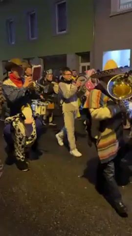 A nighttime street scene features a festive parade of people in costumes, playing musical instruments. The parade appears to be on a narrow street, lined with buildings. The people are dressed in a variety of colorful costumes including a cowboy, a pig, a mouse, a cow and a figure in a patchwork cloak. They play trumpets, a tuba, a sousaphone, and a drum, creating a lively musical atmosphere. The lighting is dim, with streetlights and light emanating from the buildings. The parade participants are moving forward, creating a dynamic flow through the street.