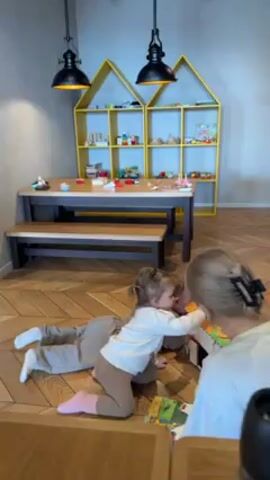 The video shows a room with a wooden herringbone floor, a table and bench, and a yellow house-shaped bookshelf with toys on it. Two young children, dressed in neutral-toned clothing, are on the floor playing with a small, house-shaped toy with a woman. The woman is also on the floor with the children, interacting with them and the toy. One child is crawling around the toy, while the other is holding a card. The woman is helping them with the toy.