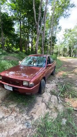 A red sedan is parked on a dirt path surrounded by trees. The car is slightly angled into the path, with its front wheels appearing to be stuck in the dirt. The car is a two-door model with a light-colored license plate. The surrounding environment is lush and green, with tall trees and vegetation. The ground is made up of dirt and some scattered leaves and debris. The sky is visible through the trees and appears to be bright.