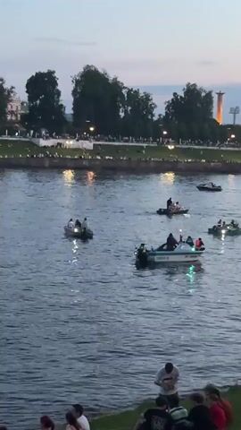 The video shows a river scene at dusk with many boats on the water. People are gathered on the grassy bank in the foreground, some sitting and others standing. Several boats, including motorboats and what appear to be smaller dinghies, are moving across the river. Some of the boats have lights illuminated. In the background, there is a treeline, a bridge with colored lights, and buildings with lights visible. The sky is a soft twilight hue with a faint crescent moon visible. The overall atmosphere suggests a leisurely evening event or gathering.