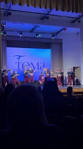 A performance by the "Tema" acrobatic dance club is taking place on a stage. The dancers, dressed in dark crop tops and maroon pants, move in synchronized choreography. The stage is lit with blue light, and the club's logo is displayed on a large screen in the background. An audience watches from their seats, their silhouettes visible in the foreground.