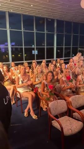 The video shows an indoor setting with a large group of young women who appear to be contestants in a beauty pageant. They are sitting in chairs, dressed in cocktail attire, and holding roses. Many of them are wearing sashes that say "Finalist".  The room has large windows offering a view of what seems to be a city at dusk. There is a disco ball hanging from the ceiling. A woman walks past the contestants, seemingly to congratulate them, while other individuals in black jackets with "UNIQUE" logos are also moving around in the foreground. The group is applauding, creating a celebratory atmosphere.
