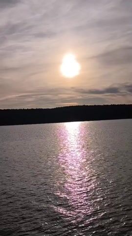 The video shows a wide shot of a body of water with a dark treeline in the background. The sun is visible in the sky, low on the horizon, casting a bright reflection on the water. The water is choppy with small waves. The sky is cloudy with a mix of light and dark clouds.