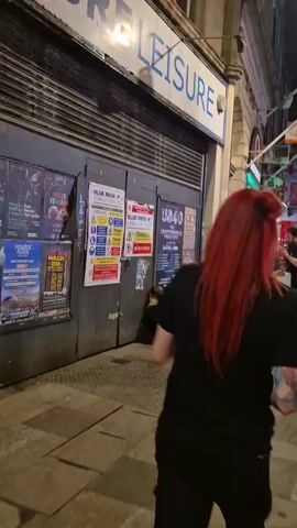 The video shows a man in shorts and a dark t-shirt dancing on a city sidewalk in front of a homeless man who is drumming. The drummer is sitting on the sidewalk with his back against the wall of a building. Several posters are attached to the wall, advertising a demolition company and a UB40 concert. The building itself has a sign that says "Leisure." Another person with red hair is visible in the corner of the frame.