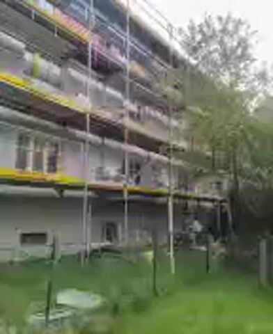 The video shows a building encased in scaffolding. The scaffolding is multi-layered, reaching up the height of the multi-story building. The building itself is a pale color, with windows visible on each level. Some trees and overgrown grass surround the base of the building. There is no visible activity. The video seems to have been taken from ground level.