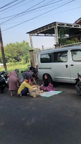 A group of four people, including two women and two men, are gathered on a paved road in front of a house with a white fence and a silver van. One woman is wearing a pink and white striped dress, and the other is wearing a black hijab and a purple patterned dress. One man is wearing a grey t-shirt and blue shorts, and the other is wearing a yellow long-sleeved shirt and blue pants. They appear to be interacting with buckets and possibly goods in a basket. Two motorcycles are parked on the left side of the road. The scene is lit by natural daylight.