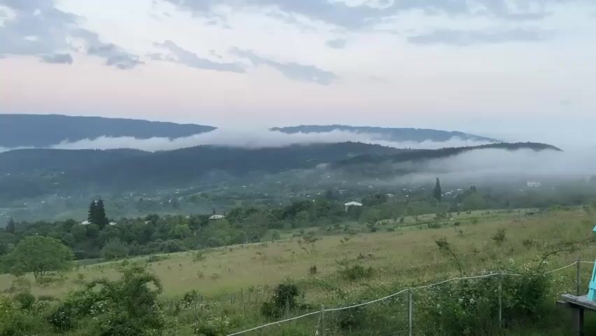 A wide shot captures a misty mountain landscape at dusk. In the foreground, a grassy field slopes gently downward, with a white rope fence running horizontally across the bottom of the frame. To the right, a bright teal swing set with a wooden platform is partially visible. In the mid-ground, a valley dotted with small houses and trees is obscured by a low-hanging layer of white fog that also clings to the forested hills beyond. The distant mountains are dark silhouettes against a pale, cloudy sky. The scene is serene and tranquil.