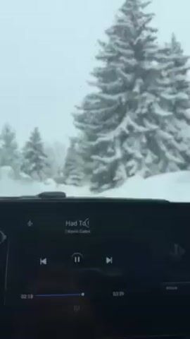 The video, shot from inside a vehicle, shows a snowy landscape. The vehicle is moving slowly along a road heavily lined with snow, with snow-covered evergreen trees visible. Inside the car, the dashboard screen displays a music player, showing the song "Had To" by Kevin Gates is playing. The weather is overcast, contributing to the snowy, wintery ambiance.