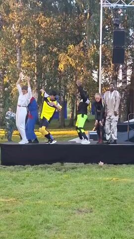 Five individuals are performing on a raised black stage outdoors. From left to right, the first person wears a white jumpsuit. The second person is in a blue and red outfit. The third person is in a yellow and black sports-like uniform with yellow socks. The fourth person wears a black top with neon green shorts and black and white sneakers. The fifth person wears a light brown, possibly metallic, patterned outfit. The stage is set in a grassy area with trees in the background. The sun appears to be setting, casting a warm light. One person on the far left is partially visible and appears to be a spectator.