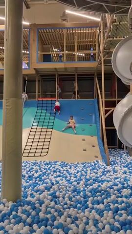 The video shows a busy indoor playground filled with children. In the foreground, a large ball pit filled with blue and white balls dominates the lower half of the screen. Several children are visible within the ball pit, playing and moving around. Towards the center of the frame, a large, multi-colored climbing wall with a net is set up. A child in a white shirt and red shorts is actively climbing the net portion of the wall. Another child in a yellow shirt is sitting in the ball pit near the base of the climbing wall, appearing to watch. In the background, there are multi-level play structures made of wood and netting, with more children and adults visible on various platforms and walkways. To the right side of the frame, large, white, circular structures, possibly slides or tunnels, are partially visible. The overall atmosphere appears to be lively and active.