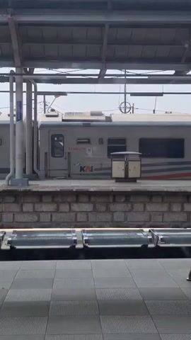 From a train station interior, a stationary train is seen through a window. The train, marked “EKSEKUTIF 5” and “KAI”, has a gray and red stripe along the side. Between the train and the camera are several rows of empty, shiny metal seats with backs. The floor consists of square gray tiles. A brick wall is along the train platform. A metal pillar stands near the train. The day appears to be bright.