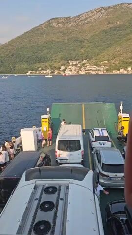 The video shows a ferry carrying vehicles and people across a body of water. In the foreground, several cars are parked on the deck, including a white van and a white car with blue stripes on its roof. Passengers are standing around the vehicles, some looking out at the water and others interacting with each other. The ferry is surrounded by calm blue water, with green, tree-covered mountains rising in the background. A few boats are visible in the distance. The sky is clear and blue with a few wispy clouds. The overall impression is a peaceful journey across a scenic waterway.