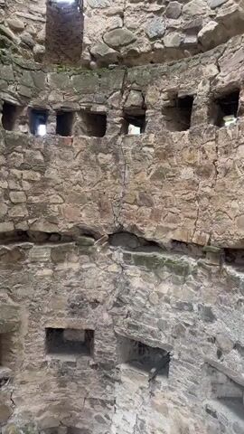 The video shows a group of people exploring an ancient stone structure. The first few seconds show the interior of the structure, with rough stone walls and small openings. Then, the view shifts to people walking on stone steps and a pathway outside the structure. The final frames reveal the exterior of the stone structure, which appears to be a historical site or monastery, with several buildings, tiled roofs, and a large gathering of people in the courtyard. The overall scene depicts tourists visiting a historical location.
