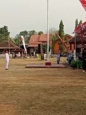 The video shows an outdoor event, likely a flag-raising ceremony, in a grassy field. A flagpole stands prominently in the center. To the left, a person in a white uniform stands at attention facing the flagpole. In the background, there are buildings with reddish-brown roofs and trees. Several other individuals are visible, some standing near a covered structure decorated with red and white banners on the right side of the frame. Flags are displayed on poles throughout the scene. The ground is covered with dry grass. The sky appears overcast.