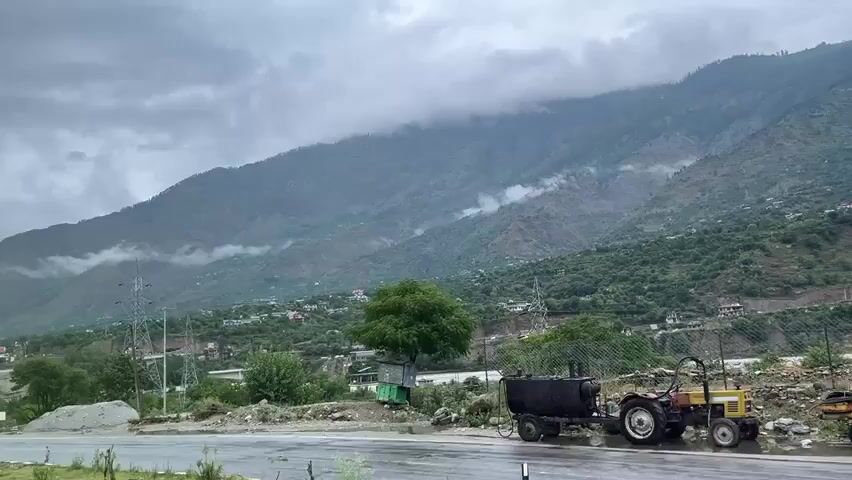 The video shows a scenic view of a mountainous landscape on a cloudy day. In the foreground, a paved road is visible, appearing wet, suggesting recent rain. To the right, a tractor with some attached equipment is parked on the side of the road. Further back, there are trees, some debris, and a small green structure. The middle ground features a river or stream flowing through a valley, with houses and trees dotting the hillsides. The background is dominated by large, verdant mountains partially obscured by low-hanging clouds, giving the scene a dramatic and somewhat misty atmosphere. Power lines and transmission towers are also visible, indicating some level of human infrastructure in the otherwise natural setting.