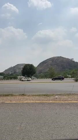 The video captures a daytime scene on a road in Kunkwaba, Nigeria, with a clear sky and scattered clouds. In the foreground, a paved road stretches horizontally across the frame, with blue and white concrete barriers separating lanes. Vehicles, including a black SUV, a grey SUV, and a black pickup truck, drive along the road. In the background, prominent hills or mountains dominate the landscape. Nestled among trees on one of the hills, a large white Ferris wheel is clearly visible, suggesting an amusement park or entertainment area. Street lights are also seen alongside the road. No specific text is visible in the video.