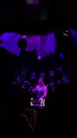 A dimly lit club scene is shown from the audience's perspective, with a DJ in the foreground playing music. The DJ, a woman with a headset, stands behind her equipment, which is illuminated. Behind her, a backdrop includes a disco ball, sheer fabrics, and a balloon garland that spells "HAPPY NEW YEAR." Purple lighting fills the scene. The silhouettes of people dancing are visible in the foreground, with one person holding a drink.