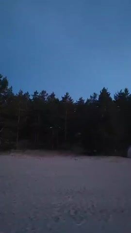 The video shows a beach scene at dusk. The sky is a deep blue, fading to lighter shades near the horizon. There are trees in the background, and a tent is visible on the sand. The water is calm, reflecting the colors of the sky. The scene transitions to a wider shot of the beach, with a faint orange glow on the horizon indicating the setting sun. The video then shows the sky as it darkens, with some clouds visible. The water is still calm, and the beach is sparsely populated with rocks. The video ends with a shot of the beach at night, with the sky completely dark and the water reflecting a faint light from the shore.