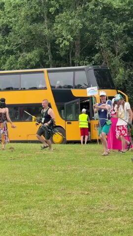 The video shows a group of people near a yellow double-decker bus on a grassy area with trees in the background. A person on the far right, seen from the back, is raising their arms in the air. To their left, a person in a white t-shirt and floral shorts is holding a pole with what appears to be a toy animal on top. Next to them, a person in a blue t-shirt and green shorts is also holding a pole with a rainbow flag. Further left, a person in a black top and a green kilt-like garment is holding two poles. To the left of them, a person in a patterned dress is walking away from the bus. Another person in a bright pink dress is visible behind the group with the poles. A person in a yellow vest is standing near the bus door. The environment appears to be outdoors during the day, likely in a field or park.