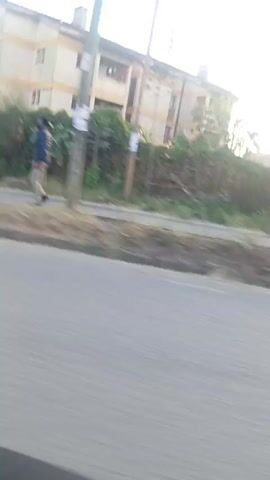 The video is taken from a moving vehicle on a road, with buildings and trees visible along the side. There are two people walking on the sidewalk beside the road. One person is wearing a blue shirt and khaki pants. The second person is wearing a black outfit. The video has bright sunlight shining through the scene. There also appears to be a bus shelter on the sidewalk in disrepair.