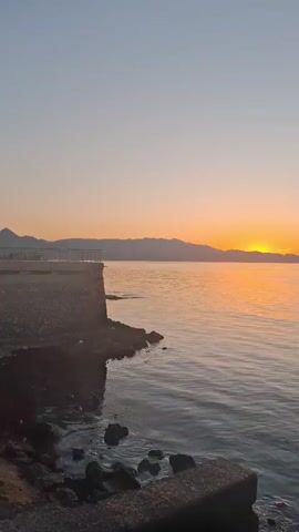 The video shows a coastal scene at sunset. The camera is positioned on a stone pier or embankment, with rocks visible along the waterline. The water is calm and reflects the orange and yellow hues of the setting sun. In the distance, a mountain range is silhouetted against the sky, which is a gradient of light blue to orange near the horizon. The sky is mostly clear, and the light from the sunset creates a warm, peaceful atmosphere. There are a few people talking in the background, but they're not visible in the shot. The scene feels serene and natural.