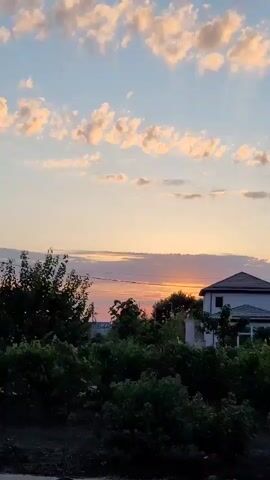The video shows a sunset over a suburban landscape. The sky is filled with pink and orange clouds against a pale blue background. In the middle ground, a white house with a dark roof is visible, surrounded by lush green trees and bushes. The foreground is darker, appearing to be a dirt or unpaved area. The sun is setting behind the trees, casting a warm glow across the scene. There are no people or animals visible in the video. The overall impression is one of peaceful rural tranquility.