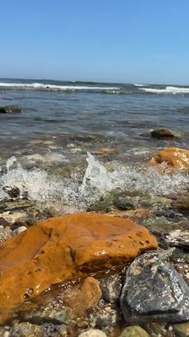 The video captures a close-up, low-angle view of the shoreline in Nea Chili, Greece, on a bright, sunny day. The foreground features several wet, smooth rocks of various sizes and colors, including prominent orange-brown, gray, white, and green hues. A large, orange-brown rock is centered in the frame, glistening with water.

Gentle waves from the sea periodically wash over the rocks, creating foamy ripples and then receding. The water is clear, revealing the colorful pebbles and stones beneath the surface. In the background, the turquoise-blue sea stretches out under a clear, light blue sky, with small waves breaking in the distance. The overall scene conveys a sense of calm and natural beauty at the beach.