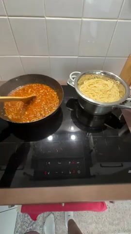 A person's feet are visible at the bottom of the frame, wearing white socks. They are standing in front of a black induction stovetop. On the stovetop, there is a black frying pan on the left, containing a red sauce with ground meat. A wooden spoon is in the pan, stirring the sauce. To the right of the pan, there is a metal colander filled with cooked spaghetti. The stovetop has a touch control panel with illuminated numbers. In the background, there is a white tiled backsplash and a wooden knife block. A red cloth is draped over the edge of the stovetop.