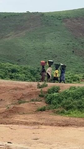 Four individuals are walking from left to right across a dusty path. They are carrying large baskets or containers on their heads. The individuals are dressed in colorful clothing, and their faces are not clearly visible. The path is bordered by lush green vegetation. In the background, a rolling green hill rises steeply. The sky appears overcast. The overall scene suggests a rural or undeveloped setting where manual labor is common.