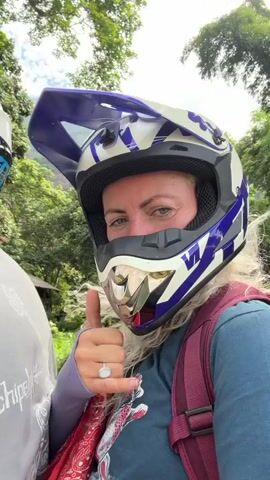 A woman wearing a blue and white motocross helmet with a silver visor smiles at the camera and gives a thumbs-up and a shaka sign. She is wearing a blue t-shirt and a burgundy backpack. Her hair is blonde and curly, and she has on a ring on her left hand. The video appears to have been filmed outdoors, with trees and greenery in the background. The camera angle is from the chest up, suggesting it is a selfie or being filmed by someone close to her.