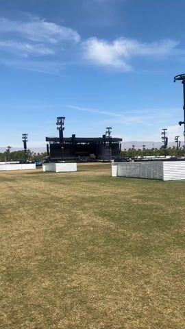 A wide-angle shot presents a Coachella stage surrounded by a sprawling, grassy field. The stage itself is a large, black structure with lighting rigs and speakers positioned on either side. Low, white barricades are scattered across the field in the foreground. In the background, palm trees and distant mountains are visible under a clear blue sky with sparse clouds. A few people can be seen moving around the stage area. The video captures the scale and setup of the festival grounds before attendees arrive.