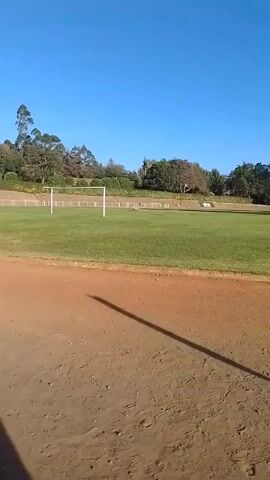 The video shows a dirt running track surrounding a grass soccer field. There are trees in the background. Three men in athletic wear, maroon and teal, are running on the dirt track. The sky is bright blue.