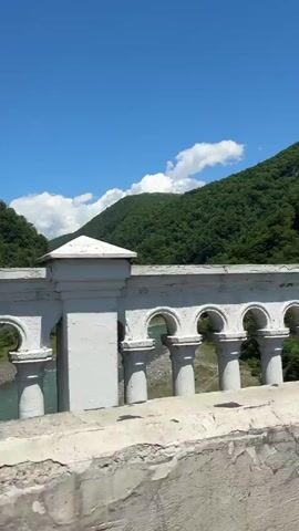 A daytime video shows a scenic view from a moving vehicle, likely crossing a bridge. The camera captures a white, ornate bridge railing with several arched openings, through which a light blue-green river is visible below. Beyond the river, lush green mountains covered in trees rise on both sides, meeting a bright blue sky dotted with white clouds. The sun is shining brightly. The sound of wind can be heard, suggesting movement. The setting is identified as Viadati Eshyra, Abkhazia. There are no people visible in the shot, nor any discernible text.